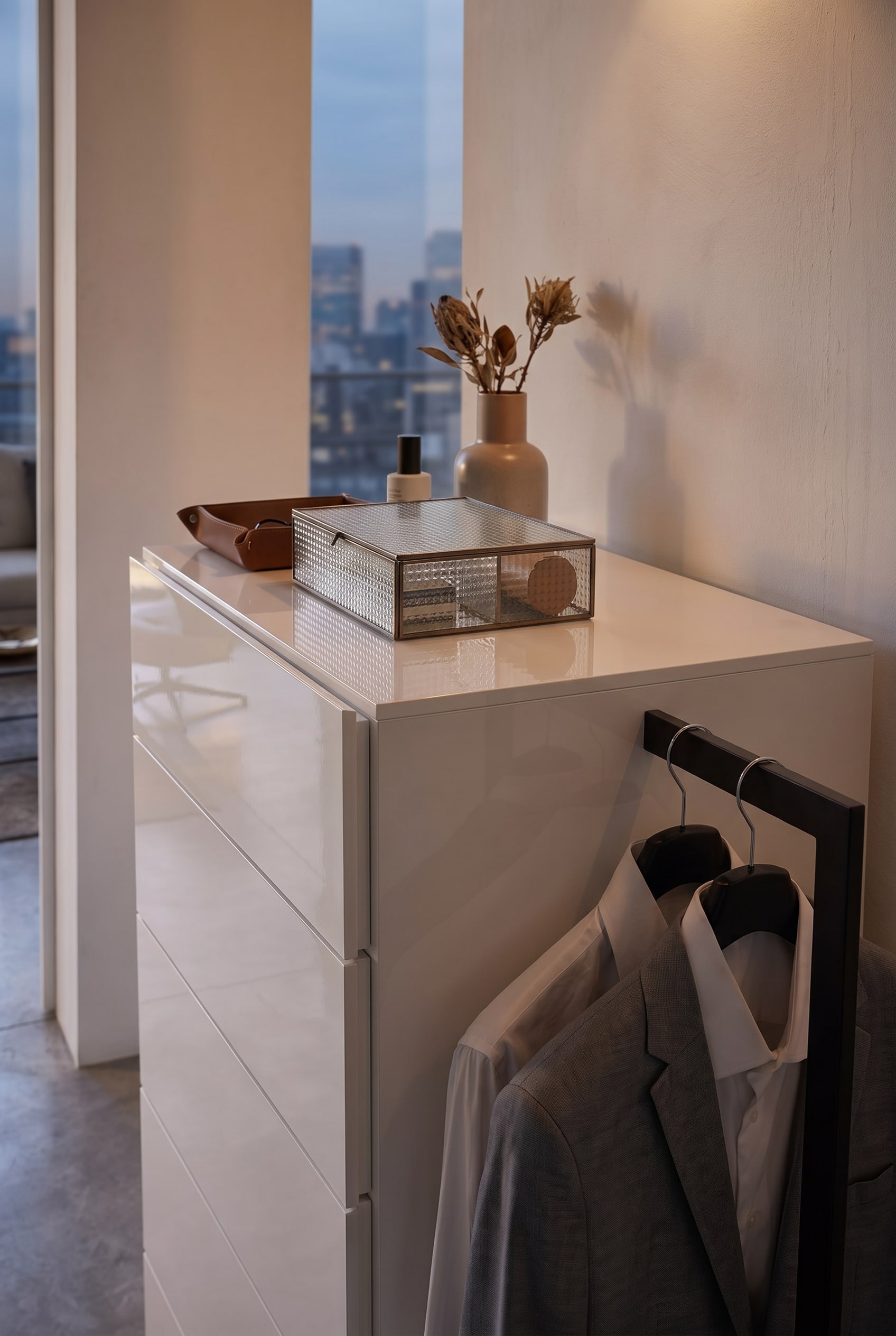 Detailaufnahme einer wei&szlig;en Kommode mit geriffelter Glasbox, Lederschale und einer seitlichen Kleiderstange mit grauem Sakko.