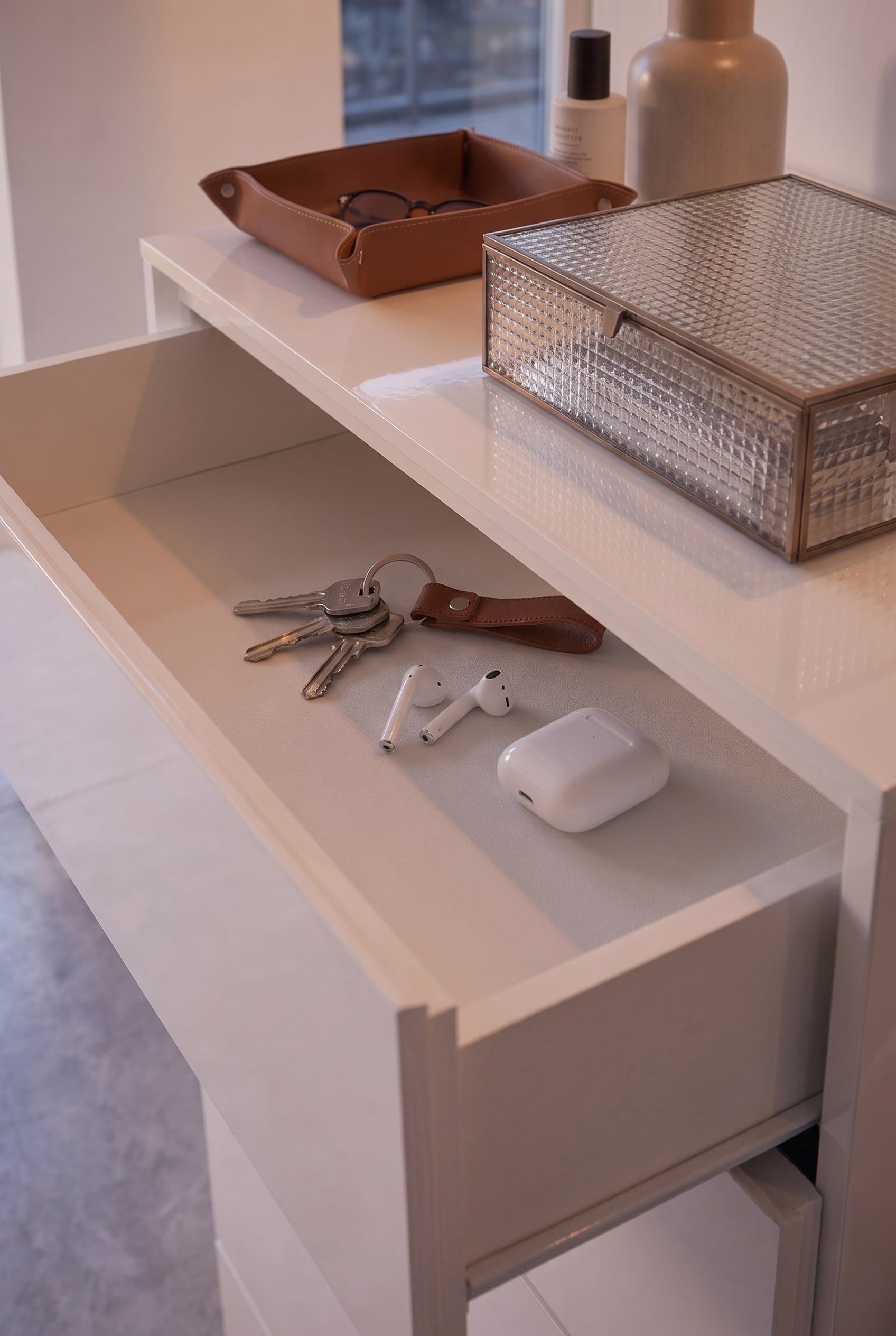 Geöffnete flache Schublade einer weißen Kommode, darin liegen geordnet Schlüssel, AirPods und ein Lederanhänger.